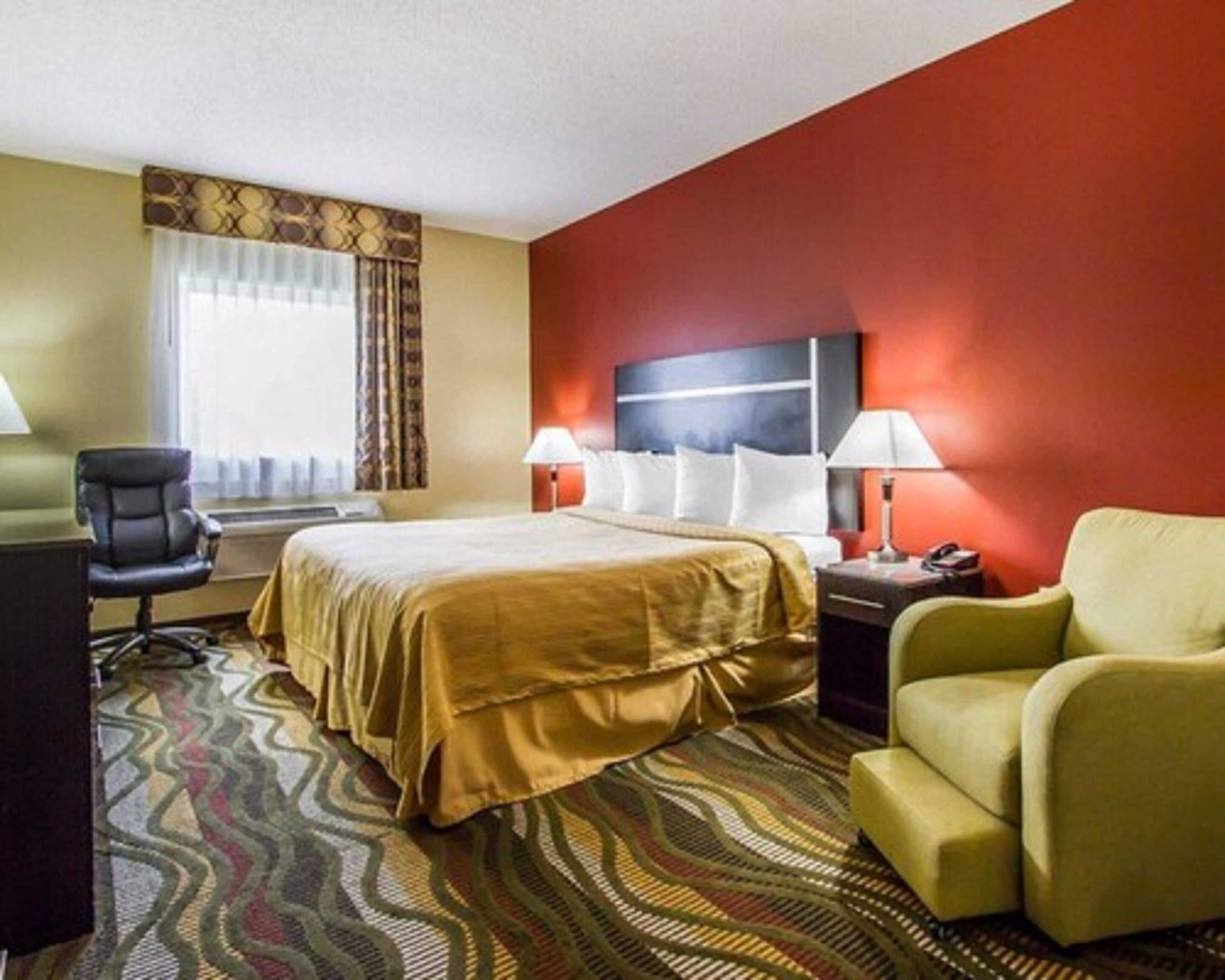 Una habitación de hotel con una cama, dos mesitas de noche con lámparas, una silla, un escritorio y una alfombra estampada.