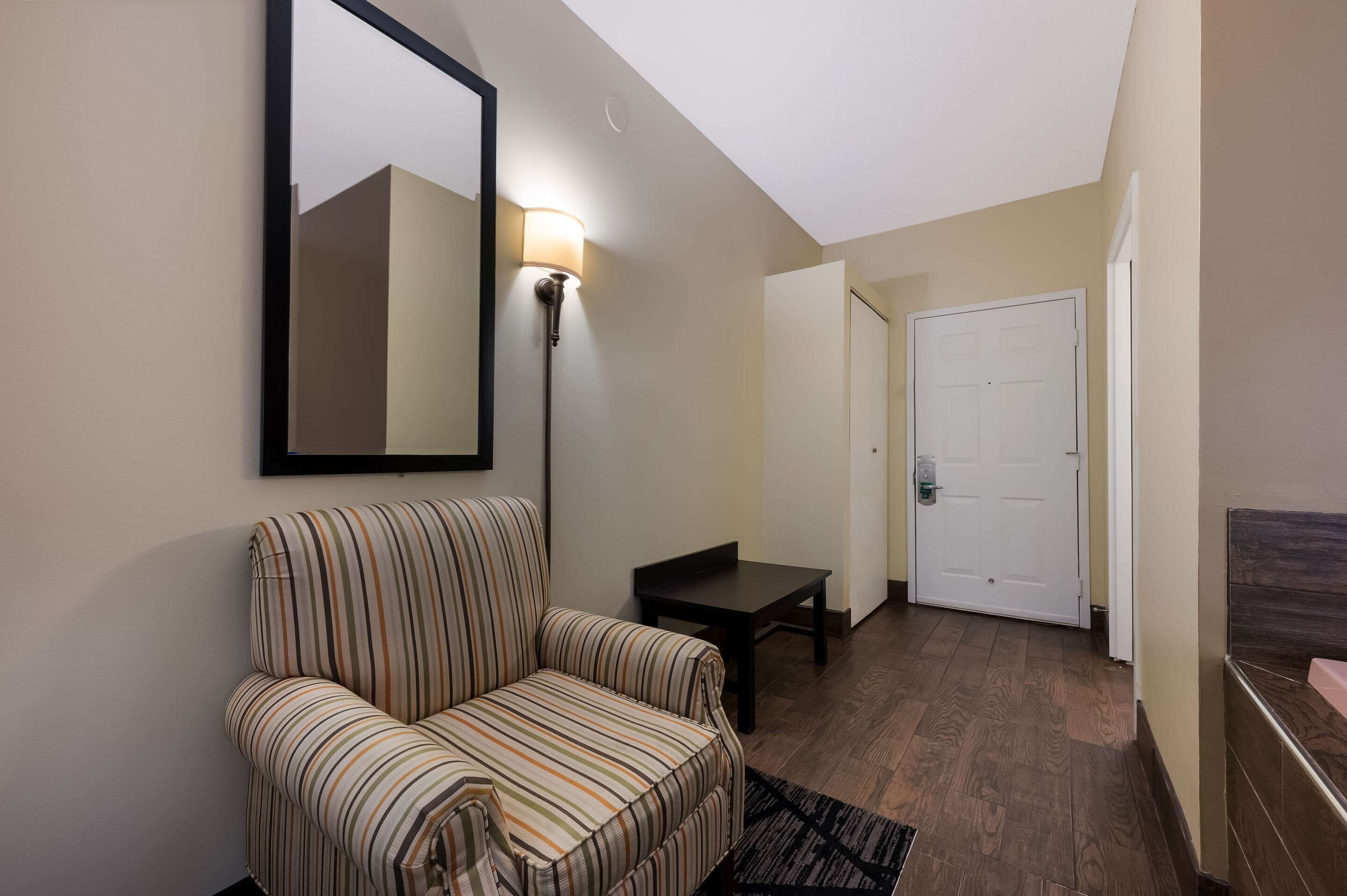 A hotel room with a striped armchair, a small table, a wall-mounted lamp, and a door in the background.