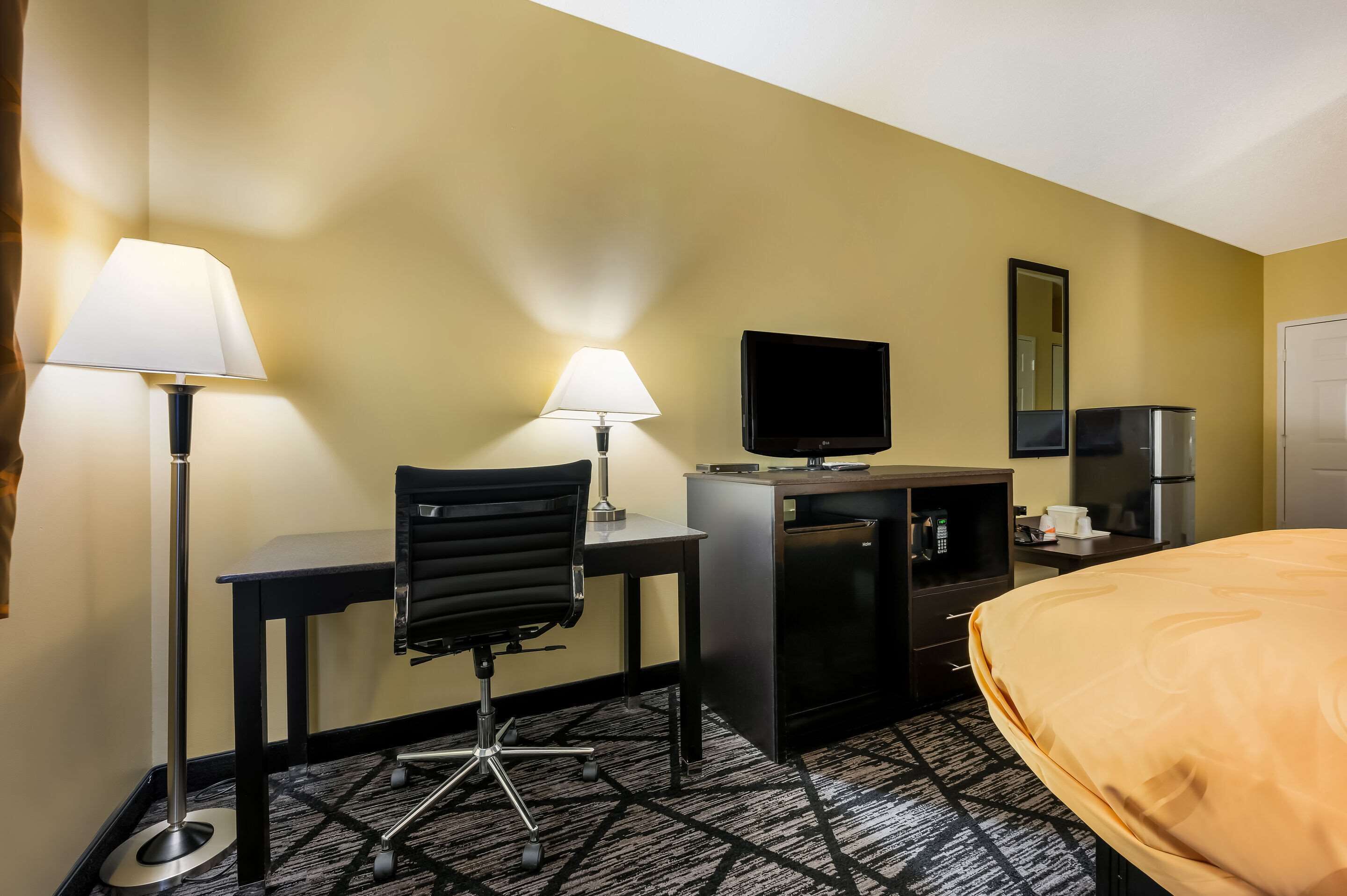 A hotel room with a desk, two lamps, a TV, a minibar, and a bed.