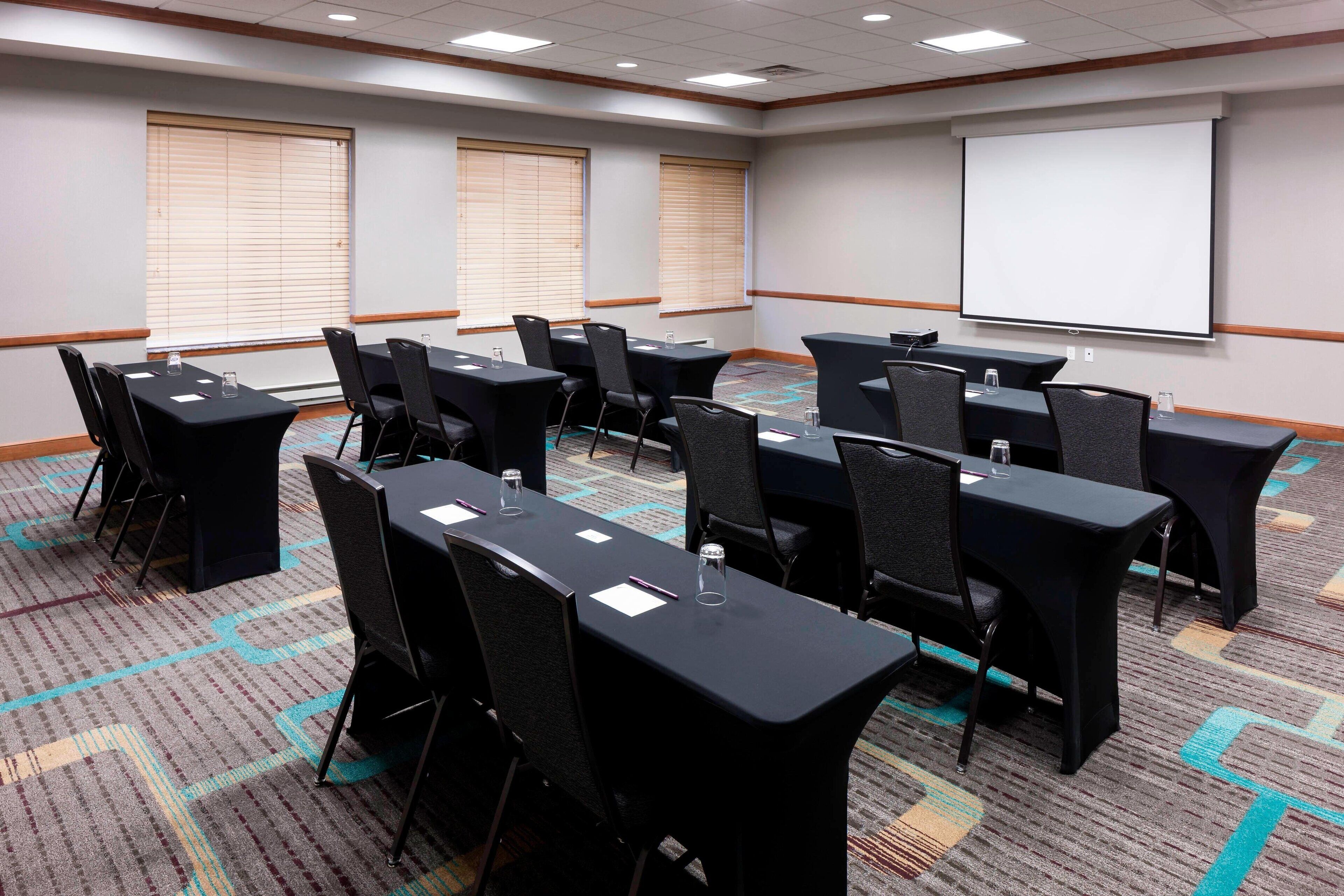 A conference room with a carpeted floor, black tables, and chairs, a projector screen, and windows with blinds.