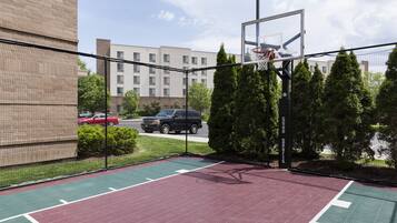 Een basketbalveld buiten met een net, omringd door een hek, en een gebouw op de achtergrond.