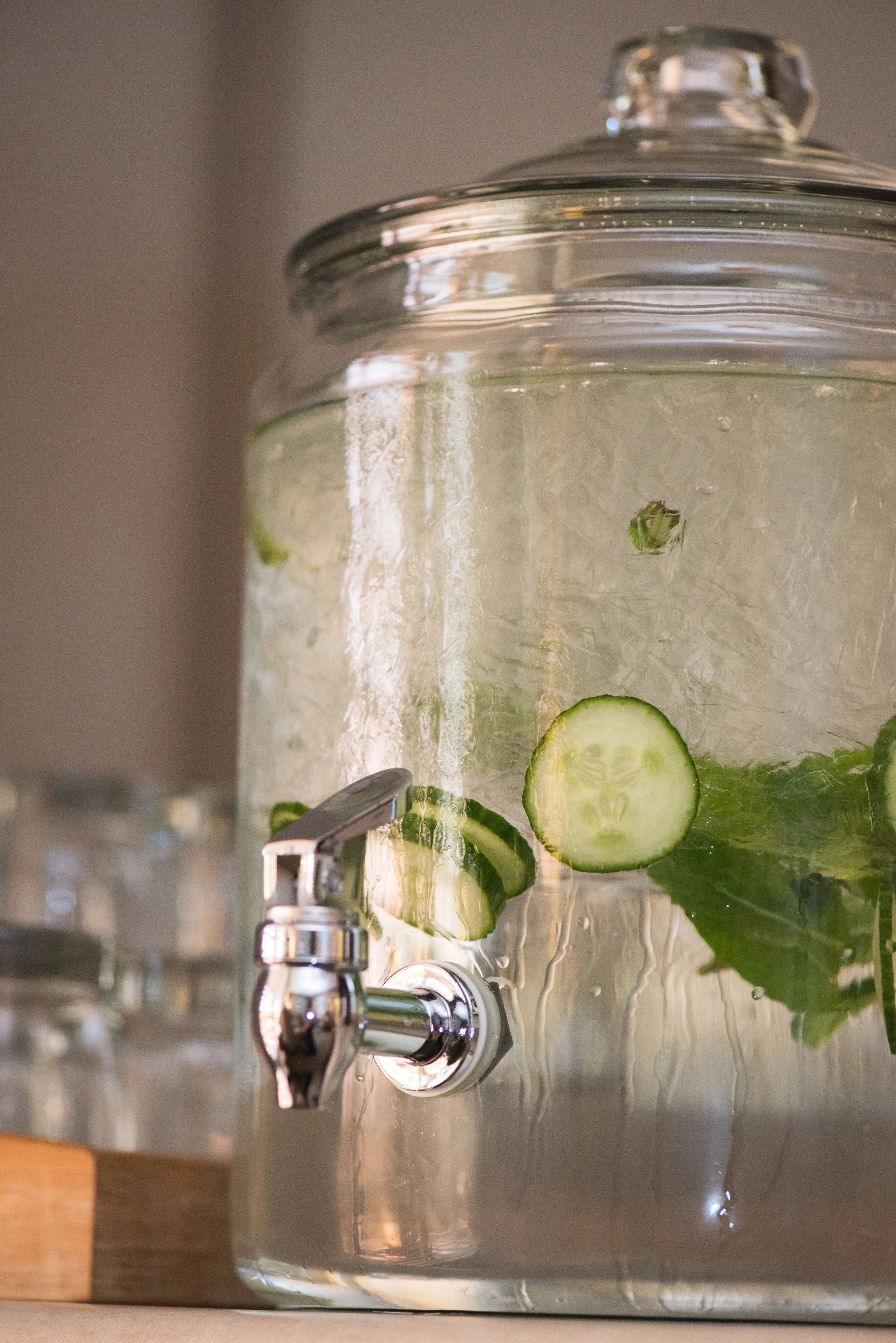 A large glass container with a tap, filled with water and slices of cucumber.