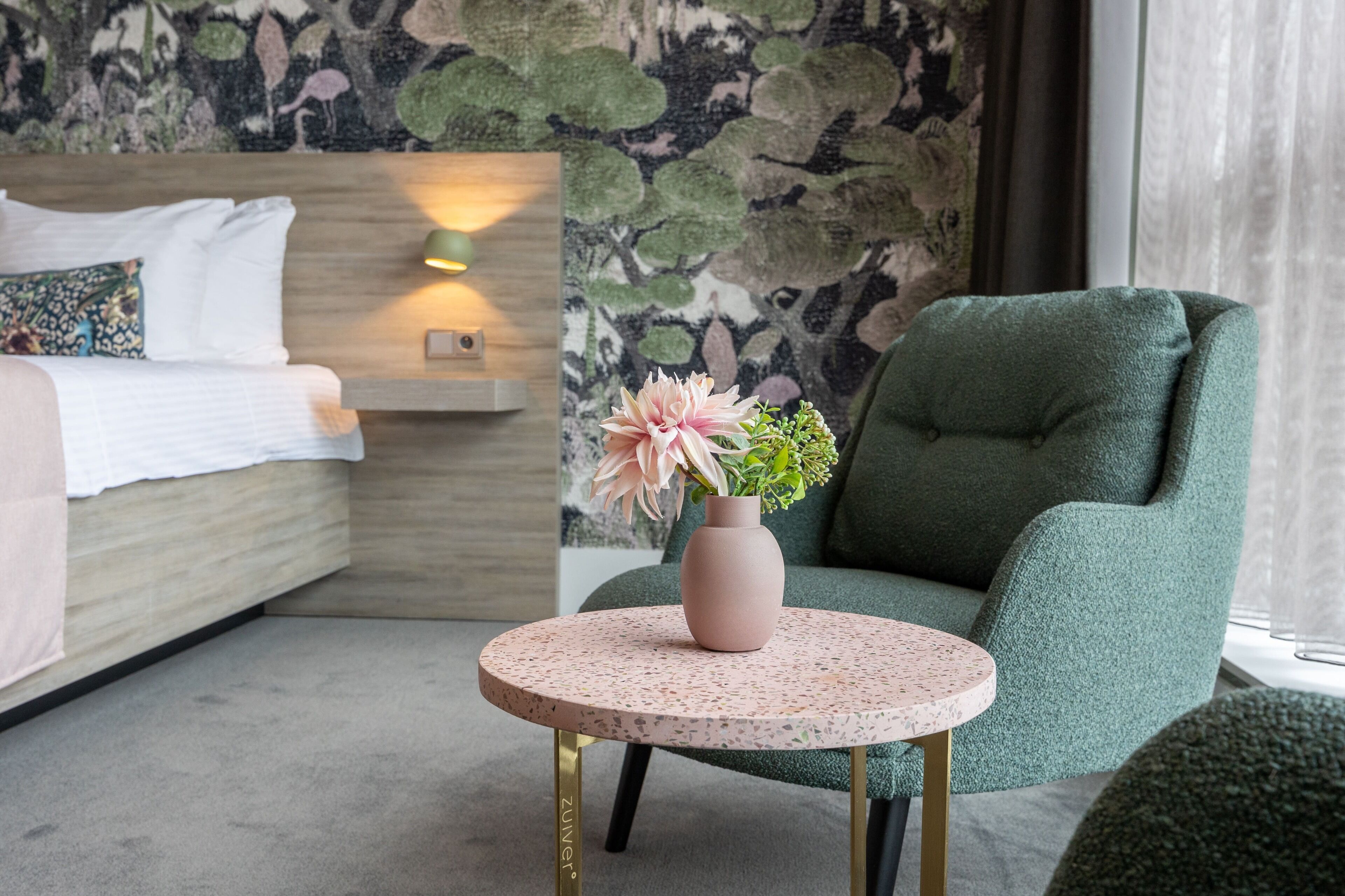 Een moderne hotelkamer met een bed, een groene fauteuil, een klein rond tafeltje met een bloevas en een muur met een bladpatroon.