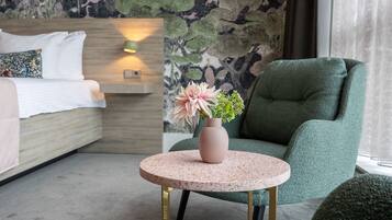 Een moderne hotelkamer met een bed, een groene fauteuil, een klein rond tafeltje met een bloevas en een muur met een bladpatroon.