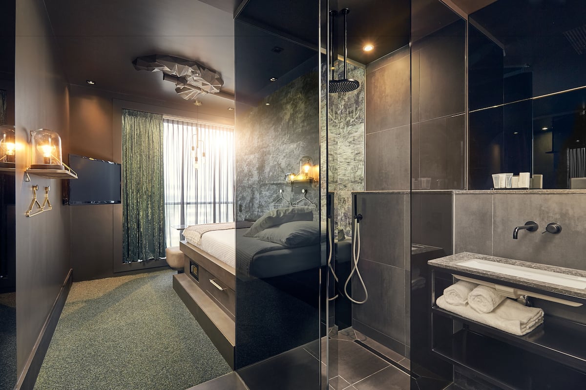 Una habitación de hotel moderna con cabina de ducha de vidrio, una cama grande, un tocador con espejo y vistas al exterior.
