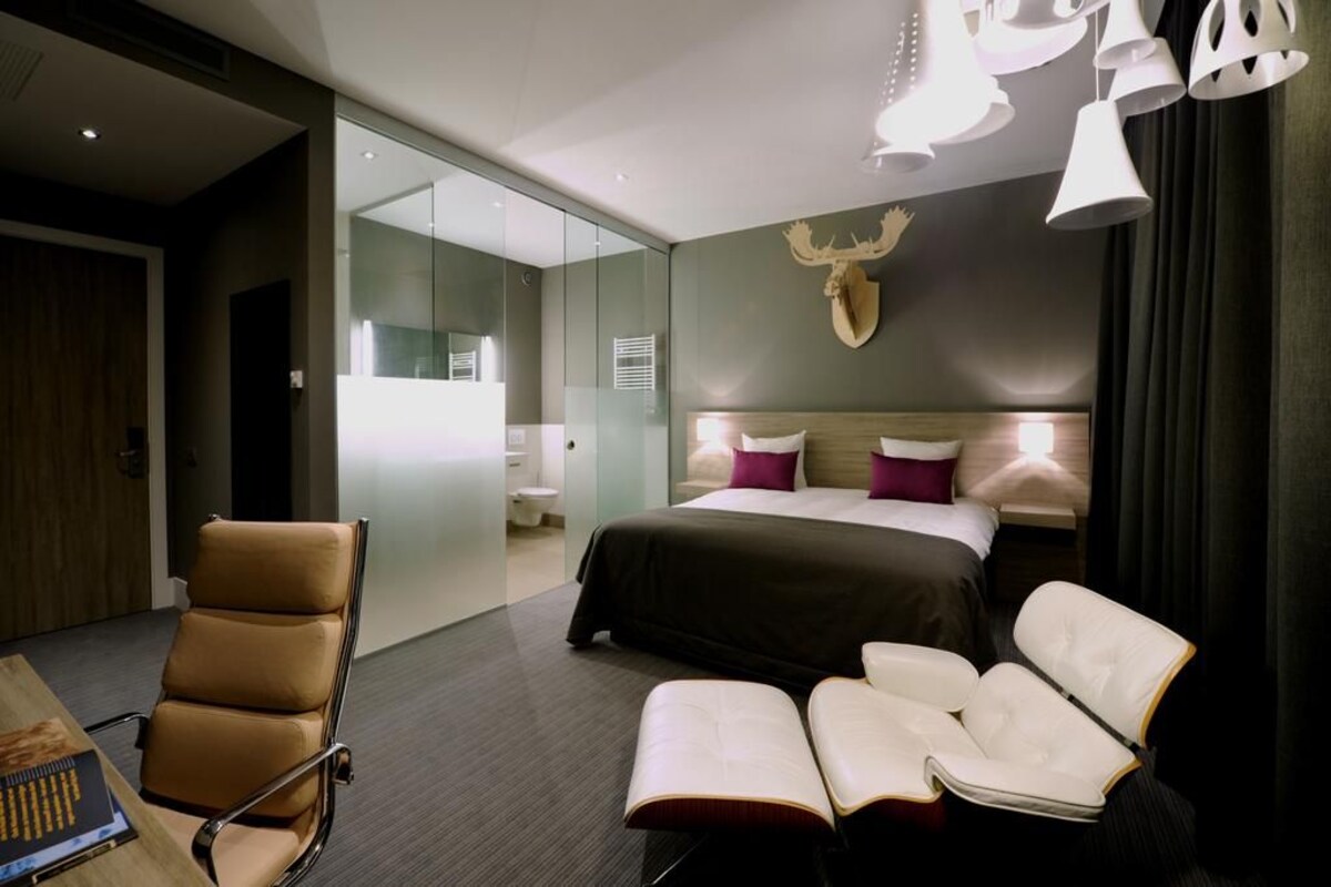 A modern hotel room with a large bed, a desk with a chair, a seating area with two chairs, and a glass partition separating the room from the bathroom.