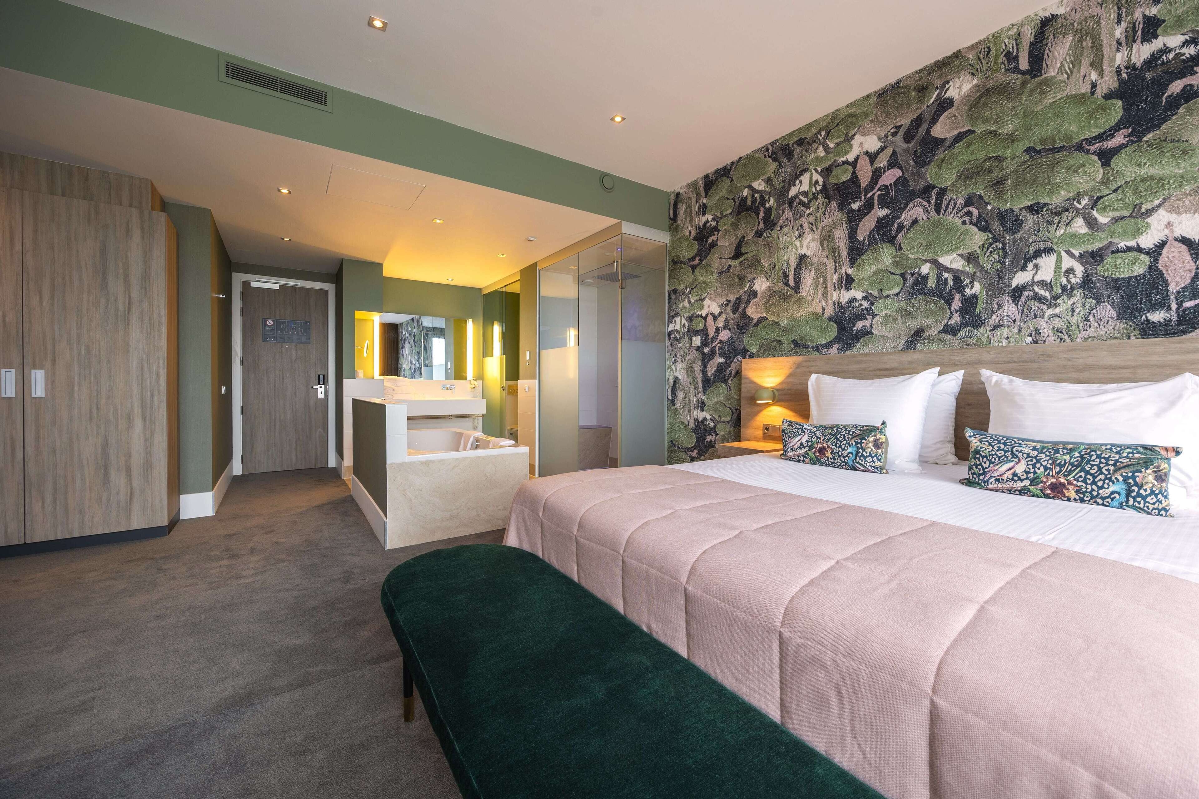A modern hotel room with a large bed, a green upholstered bench, a wooden wardrobe, a vanity with a mirror, and a mural on the wall.