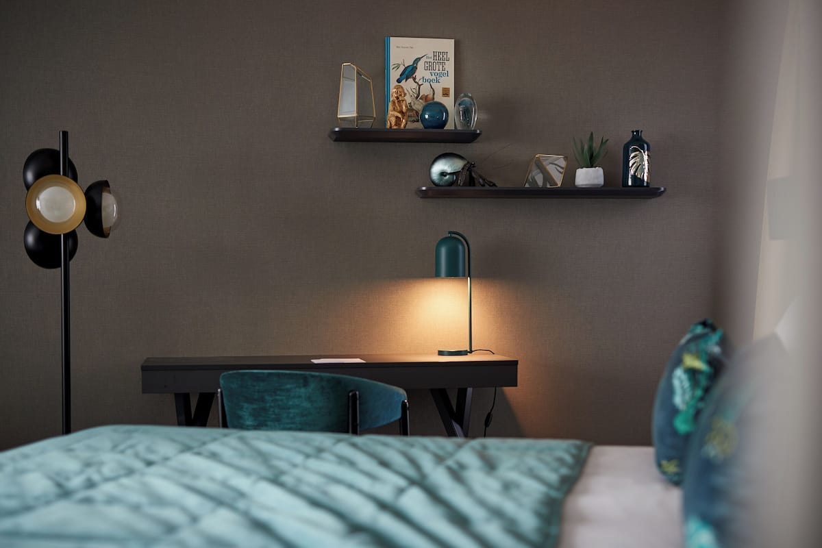 Un dormitorio con cama, escritorio, silla, lámpara y estanterías decoradas con objetos decorativos.