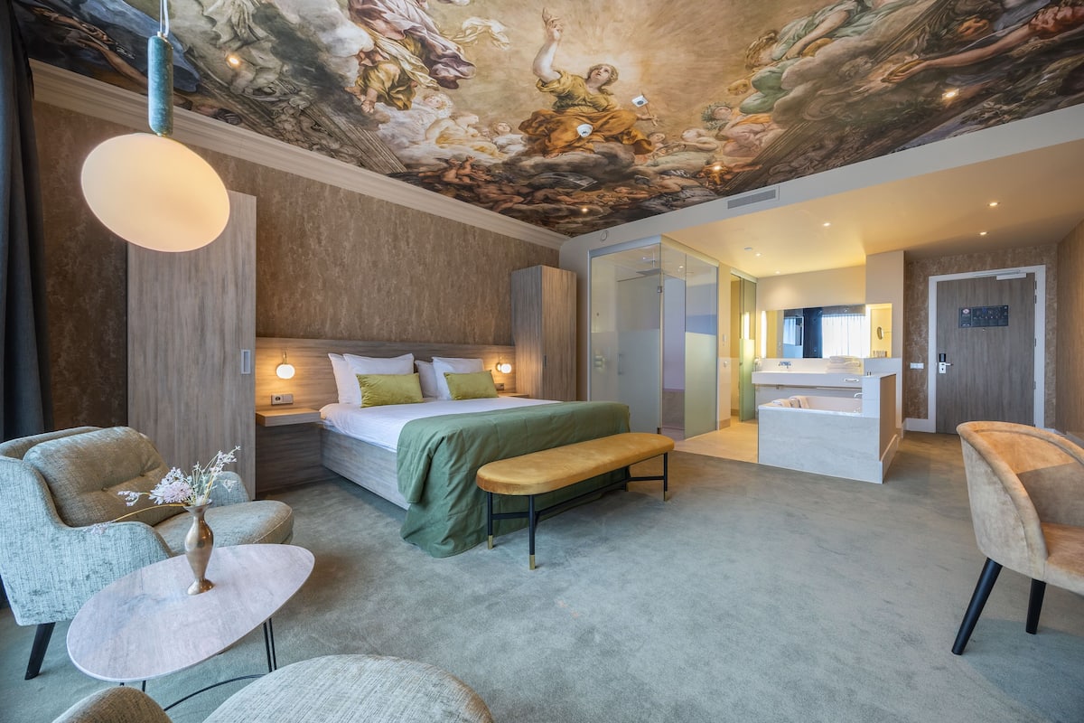 Una habitación de hotel moderna con una cama grande, un banco, una mesita con un jarrón y un mural en el techo.