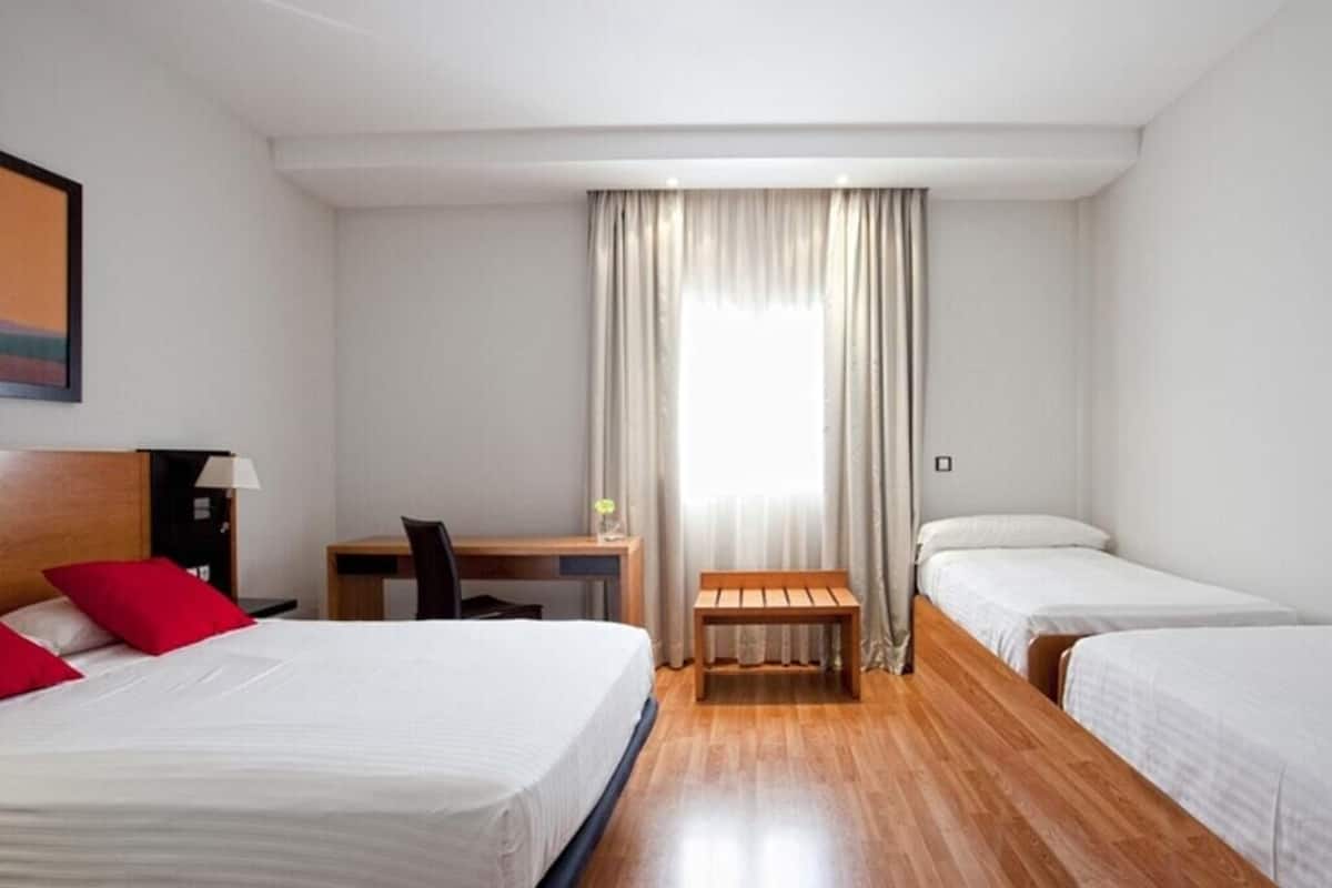 A hotel room with a bed, a desk, a chair, a bench, and two single beds.