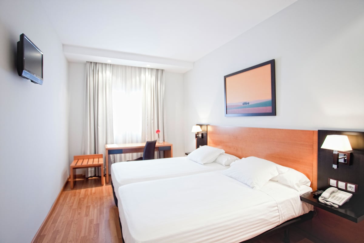 A hotel room with two beds, a desk, a chair, a TV, a lamp, and a painting on the wall.