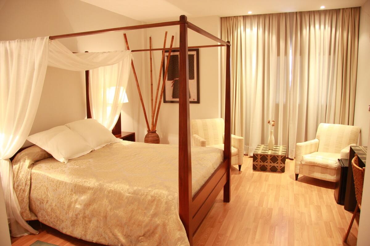 A four-poster bed with a canopy, a sitting chair, a small table with a vase, and a picture on the wall.