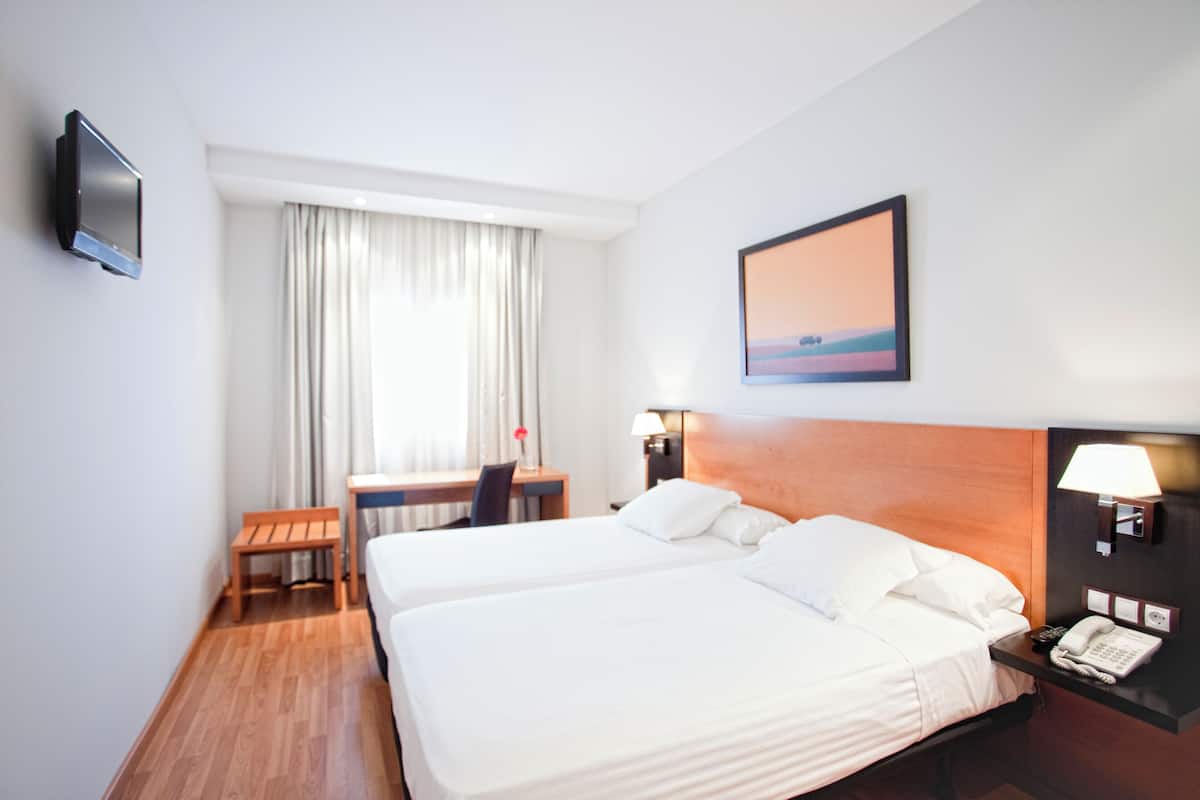 A hotel room with two beds, a desk, a chair, a TV, a lamp, and a painting on the wall.