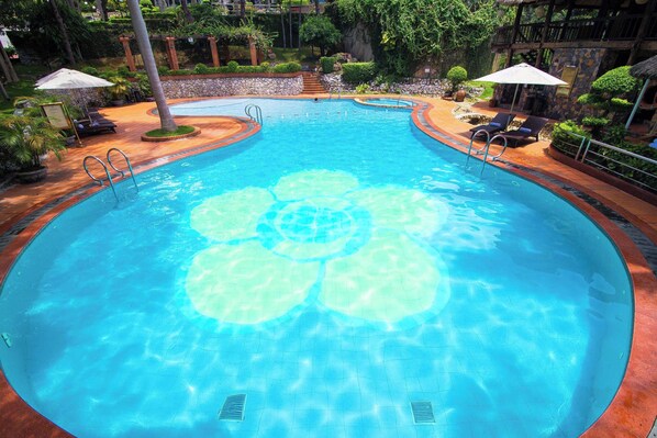 A clear swimming pool with a circular design, surrounded by lounge chairs and umbrellas, set in a landscaped area with trees and a stone wall.