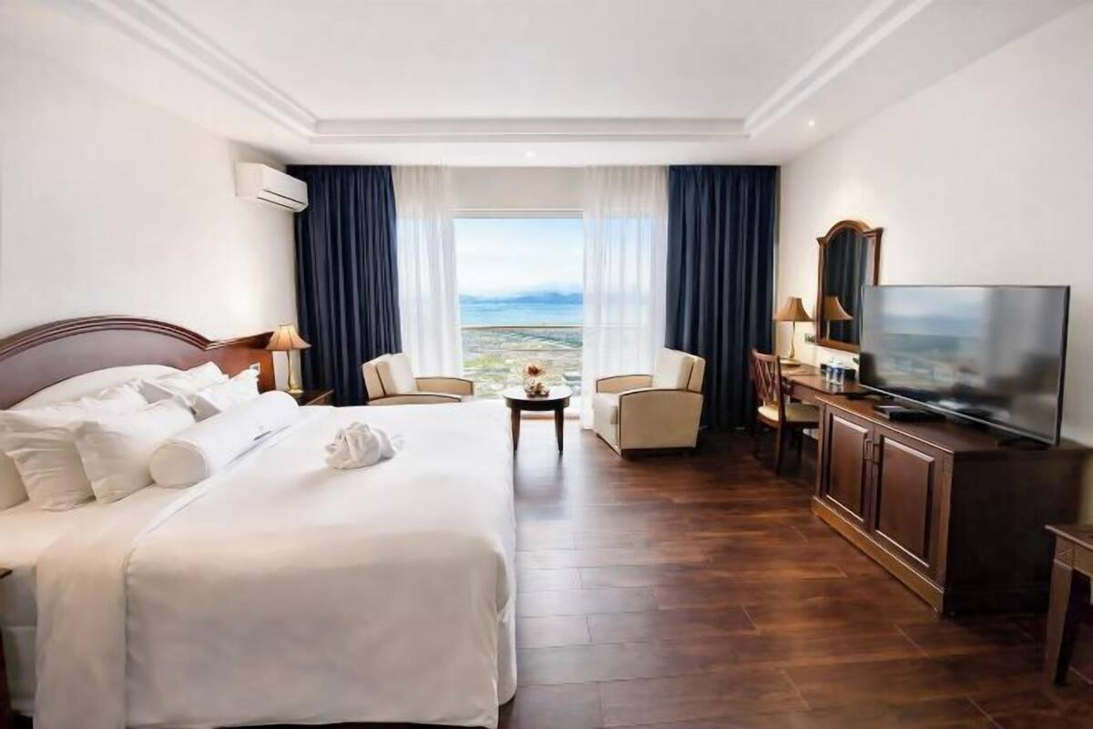 A hotel room with a large bed, a TV, two chairs, a small table, and a view of the ocean.