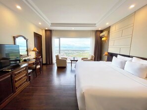 A hotel room with a large bed, a desk with a TV, a chair, a small table, and a view of the city. - Saigon Halong Hotel (Bai Chay)