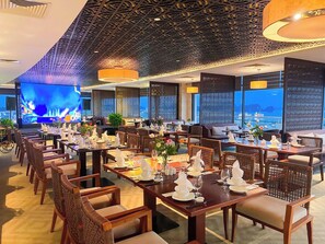A restaurant with multiple dining tables, chairs, and a large screen displaying a fire. - Saigon Halong Hotel (Bai Chay)