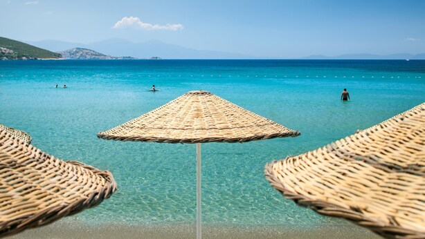 A beach with clear turquoise water, a straw umbrella, and a person in the water. - Tusan Beach Resort (Kusadasi)