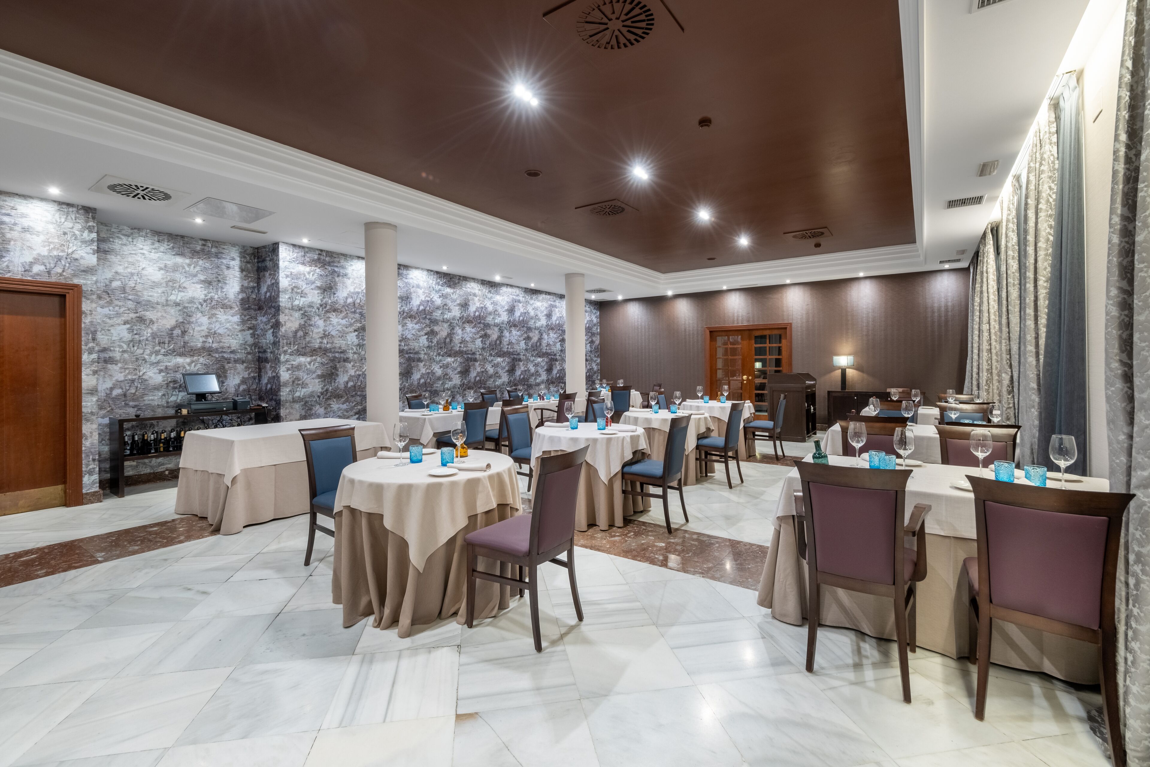Un amplio comedor con varias mesas y sillas, una zona de bar y un gran ventanal con cortinas.
