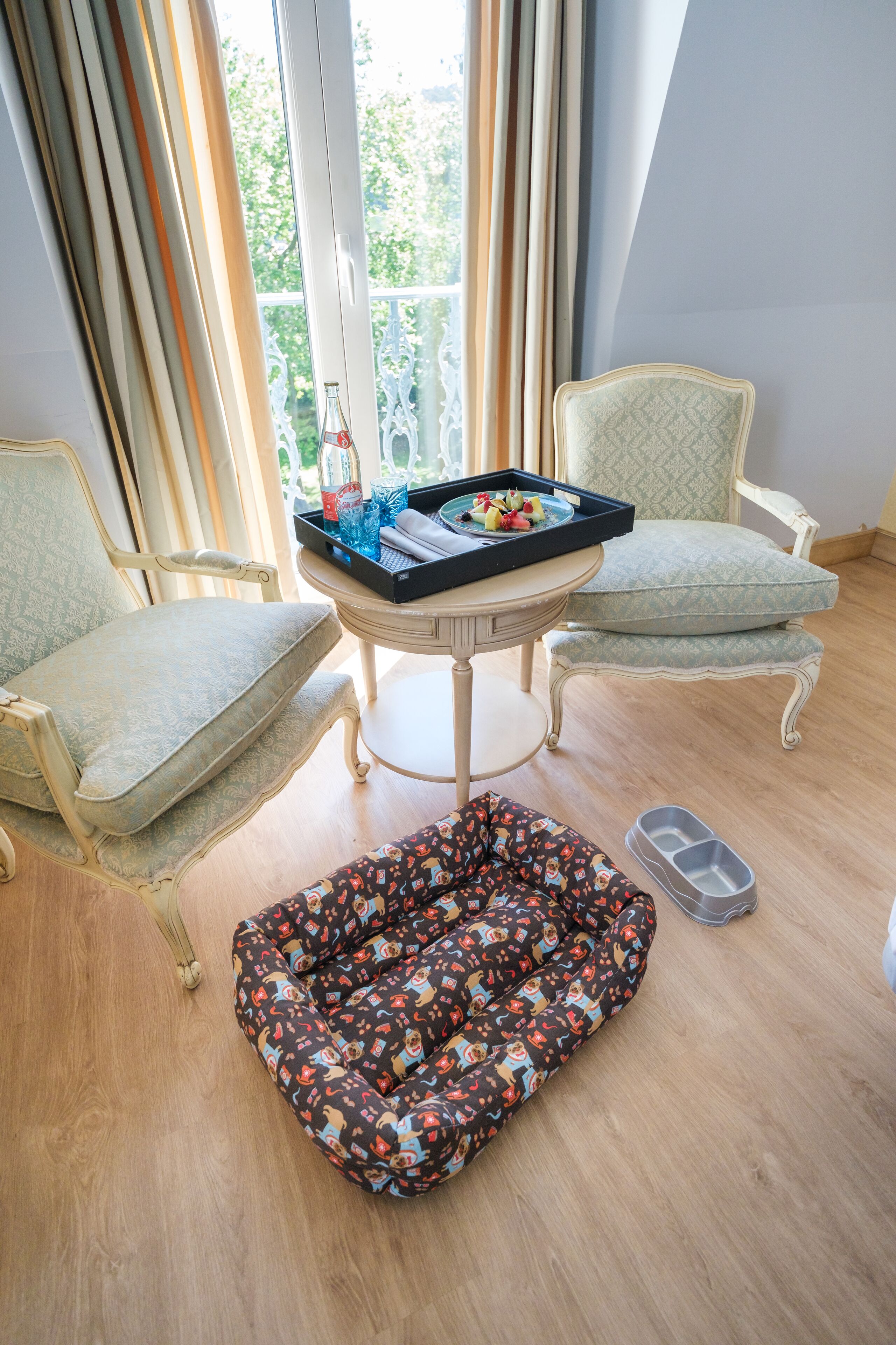 A room with two upholstered chairs, a small table with a tray of fruit and a bottle, and a patterned pet bed.