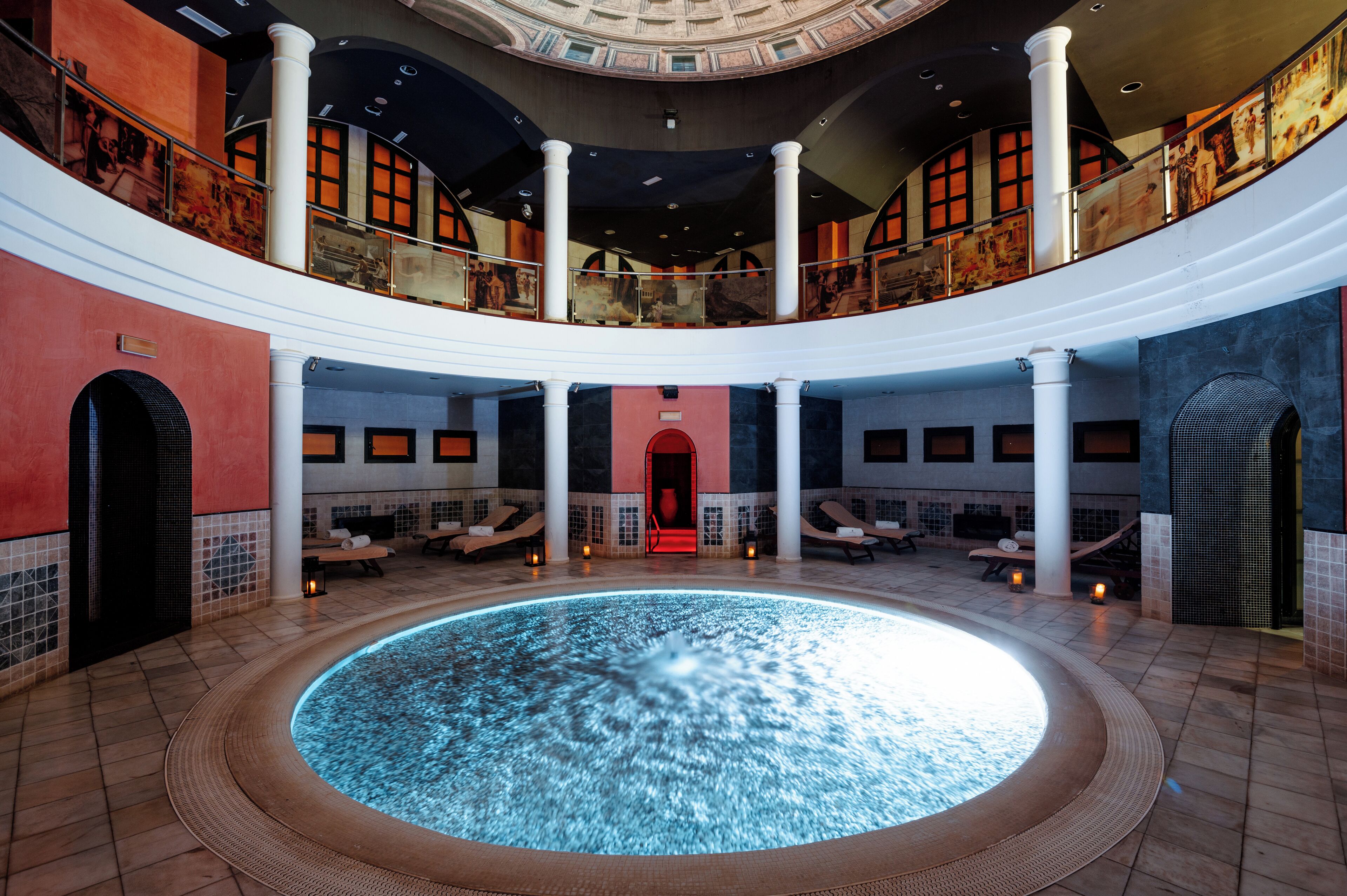 Una piscina interior amplia con una fuente central, varios espacios de descanso y un techo alto con tragaluz circular.