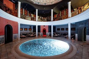Una piscina interior amplia con una fuente central, varios espacios de descanso y un techo alto con tragaluz circular.
