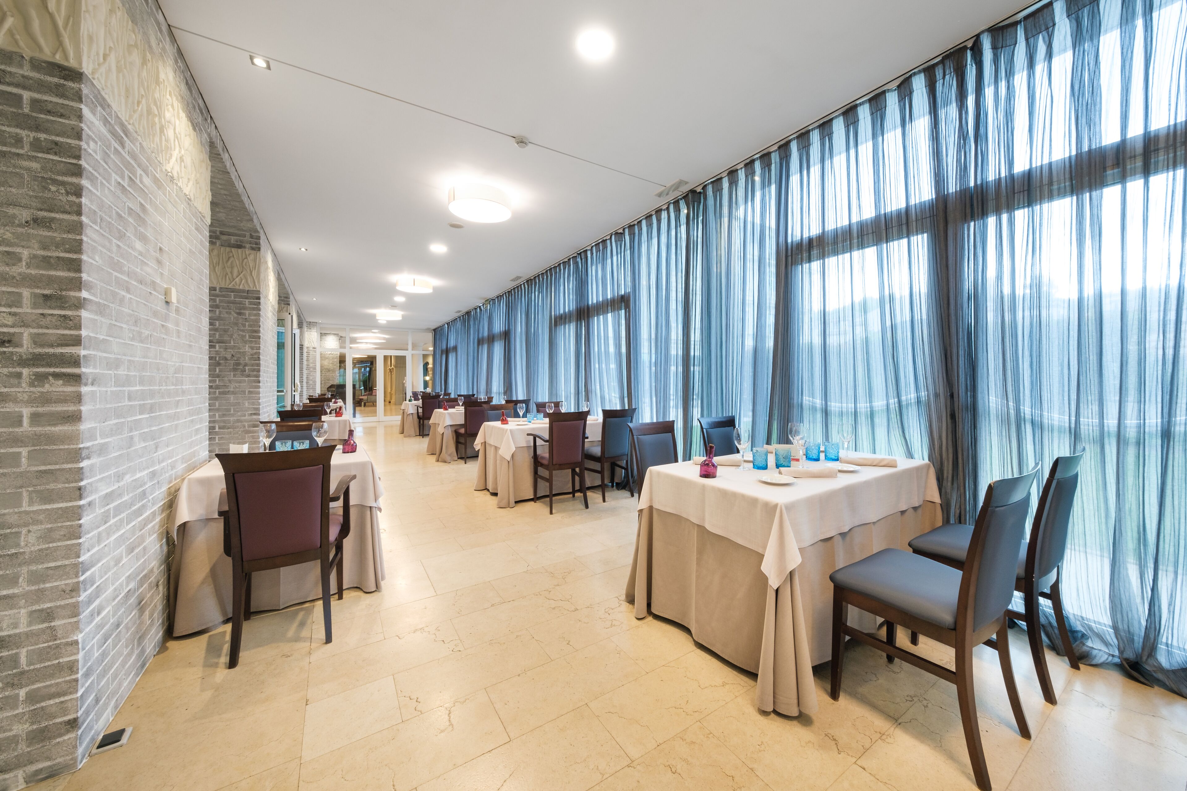 Un comedor amplio con varias mesas y sillas, ventanas grandes con cortinas azules y una pared de ladrillo en un lateral.