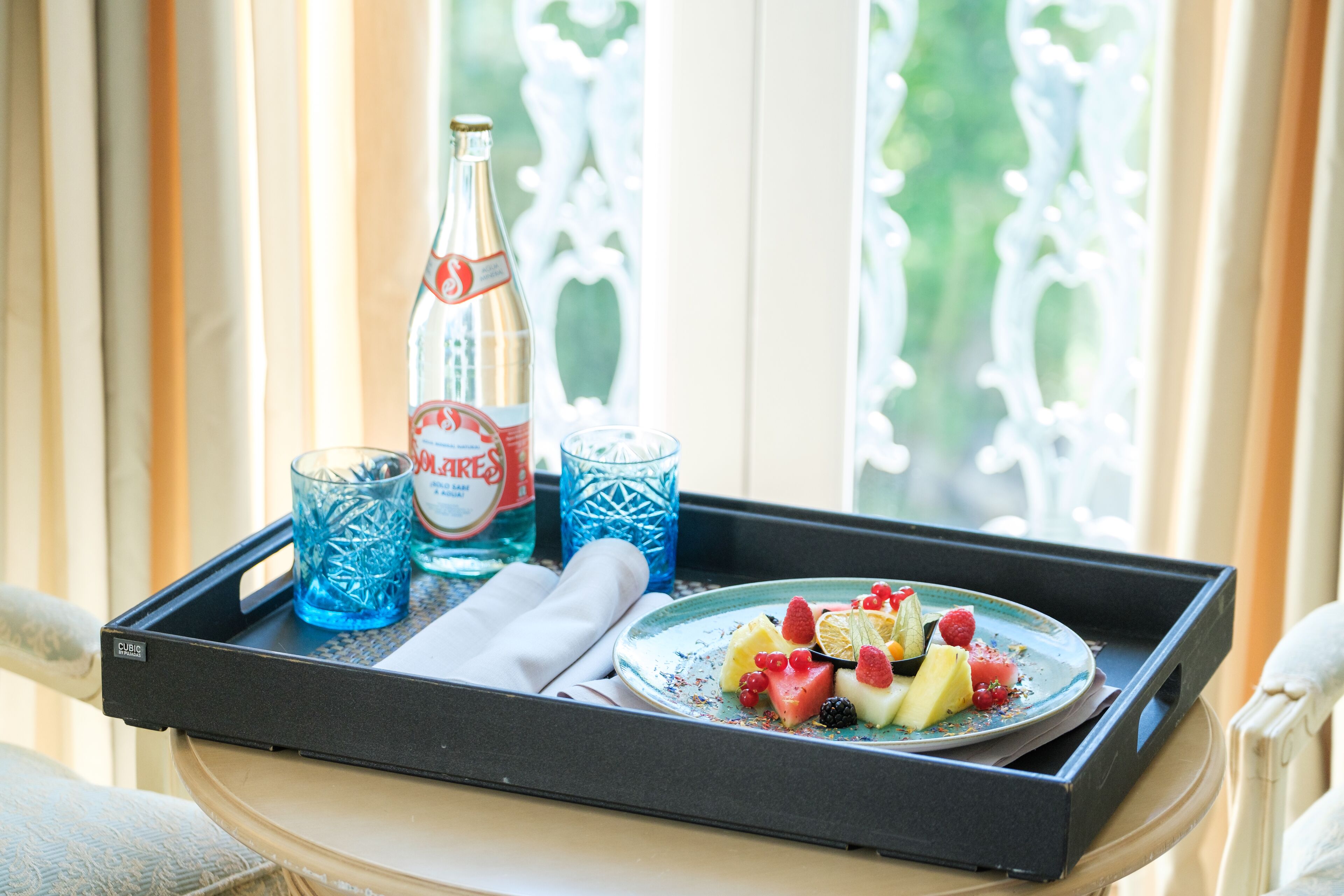 Ein Tablett mit einer Wasserflasche, zwei Gläsern, einer Obstplatte und einem Servietten auf einem Tisch neben einem Fenster.
