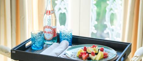 A tray with a bottle of water, two glasses, a plate of fruit, and a napkin on a table by a window.
