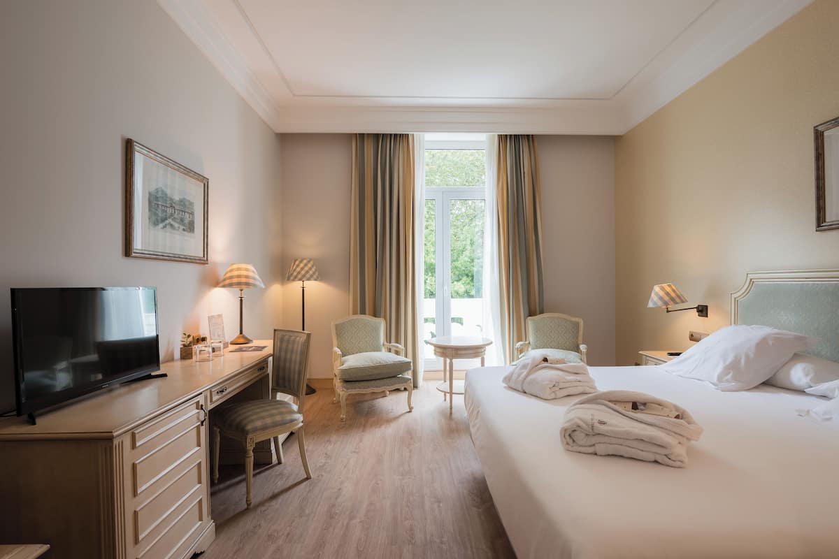 A hotel room with a bed, a desk with a TV, a chair, a small table, and a window with curtains.