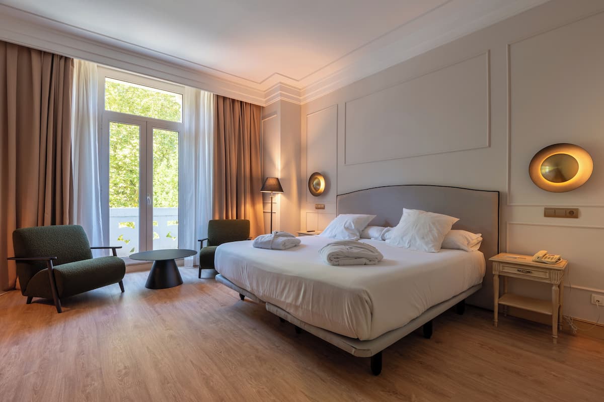 A hotel room with a large bed, two chairs, a small table, and a bedside table with a lamp.