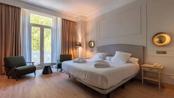 Una habitación de hotel con una cama grande, dos sillas, una mesita pequeña y una mesita de noche con lámpara.