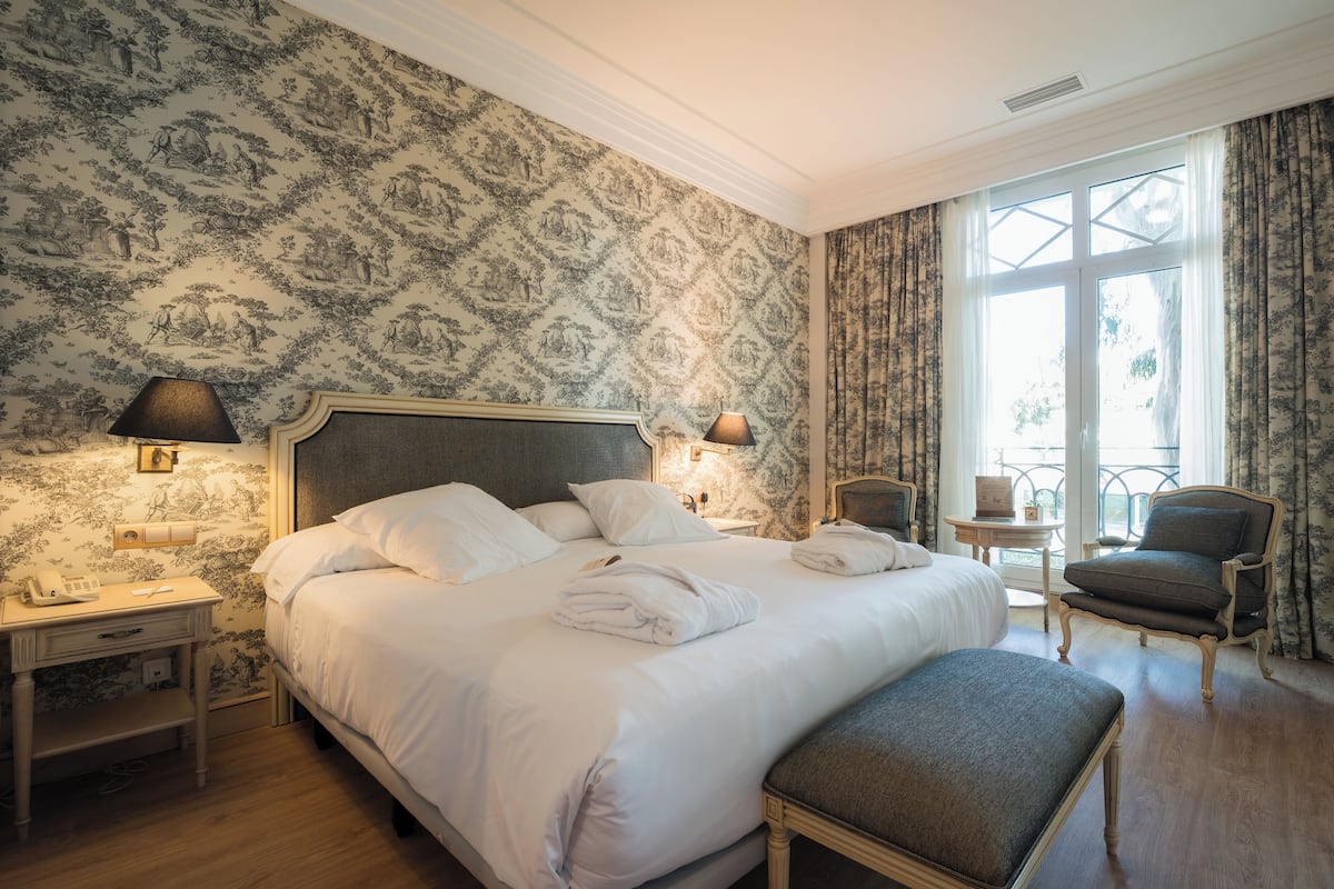 A hotel room with a large bed, two bedside tables with lamps, a chair, and a view of the outdoors through a window.