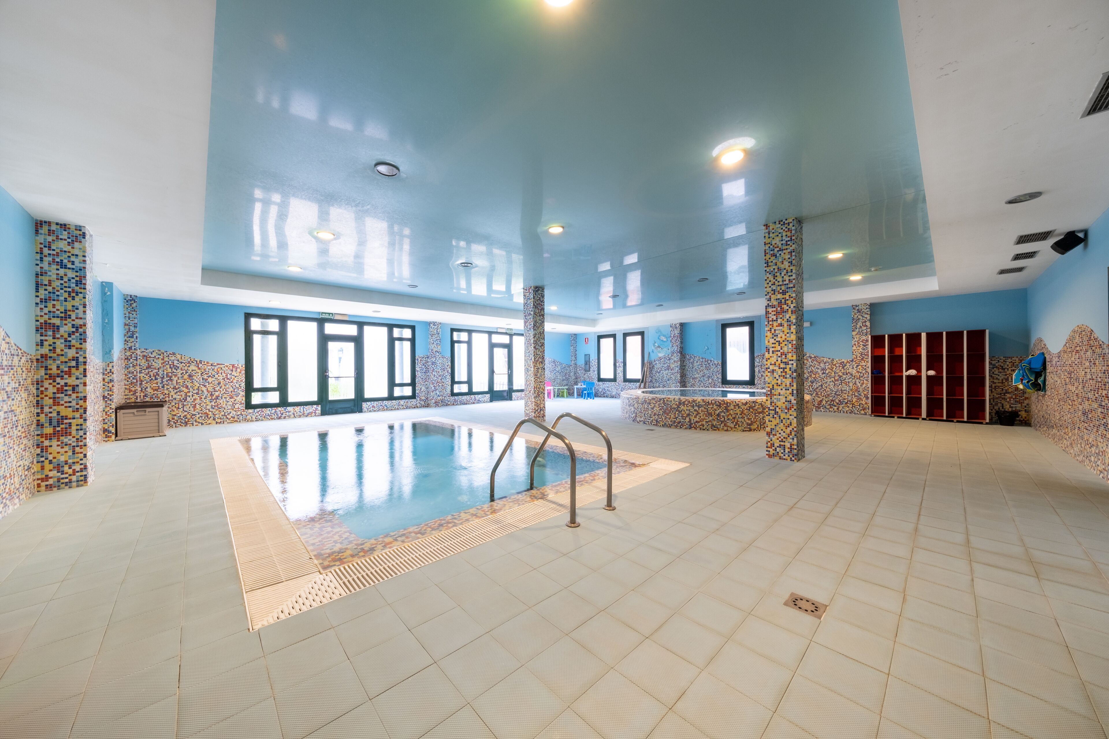 En stor indendørs swimmingpool med flisebeklædte vægge og gulv, omgivet af vinduer og søjler.