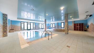 A large indoor swimming pool with tiled walls and a tiled floor, surrounded by windows and pillars.