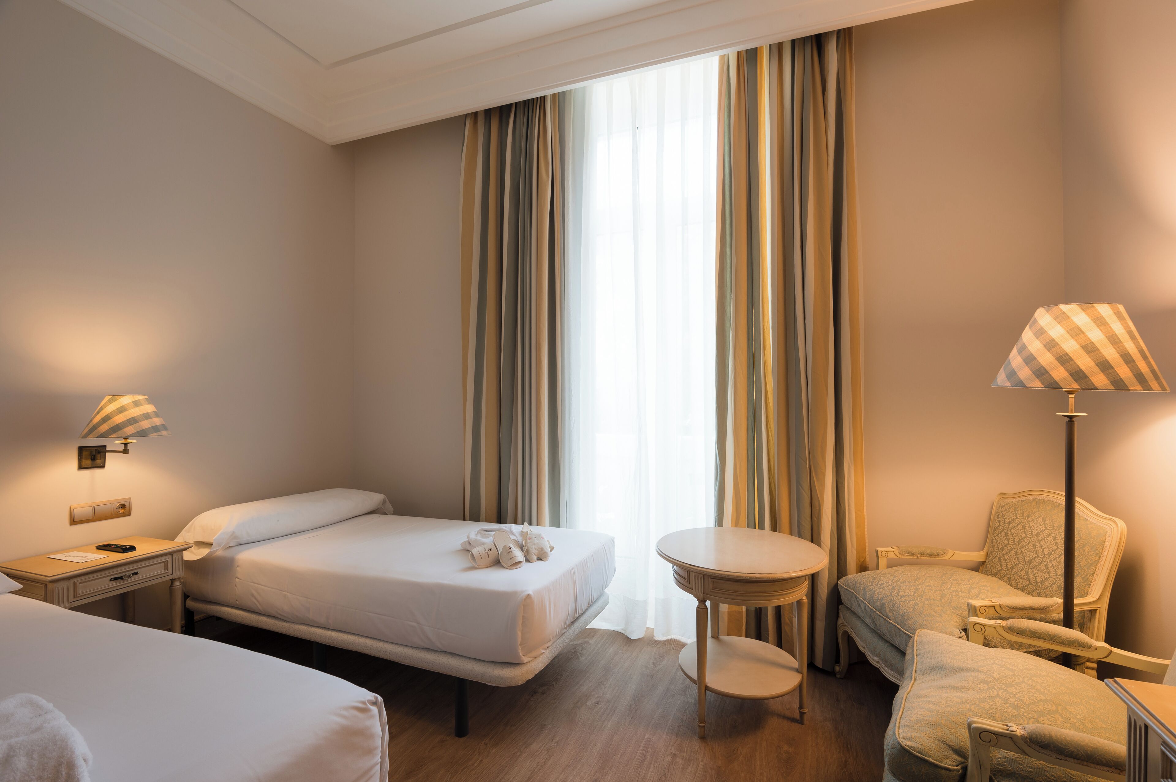 A hotel room with two beds, a small round table, a chair, and a lamp.