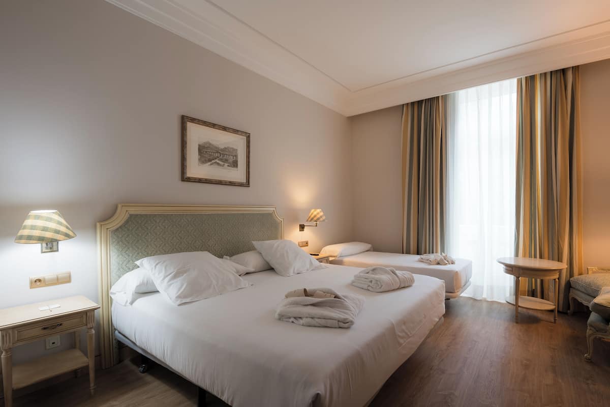 A hotel room with two beds, a bedside table, a lamp, a framed picture on the wall, and a window with curtains.