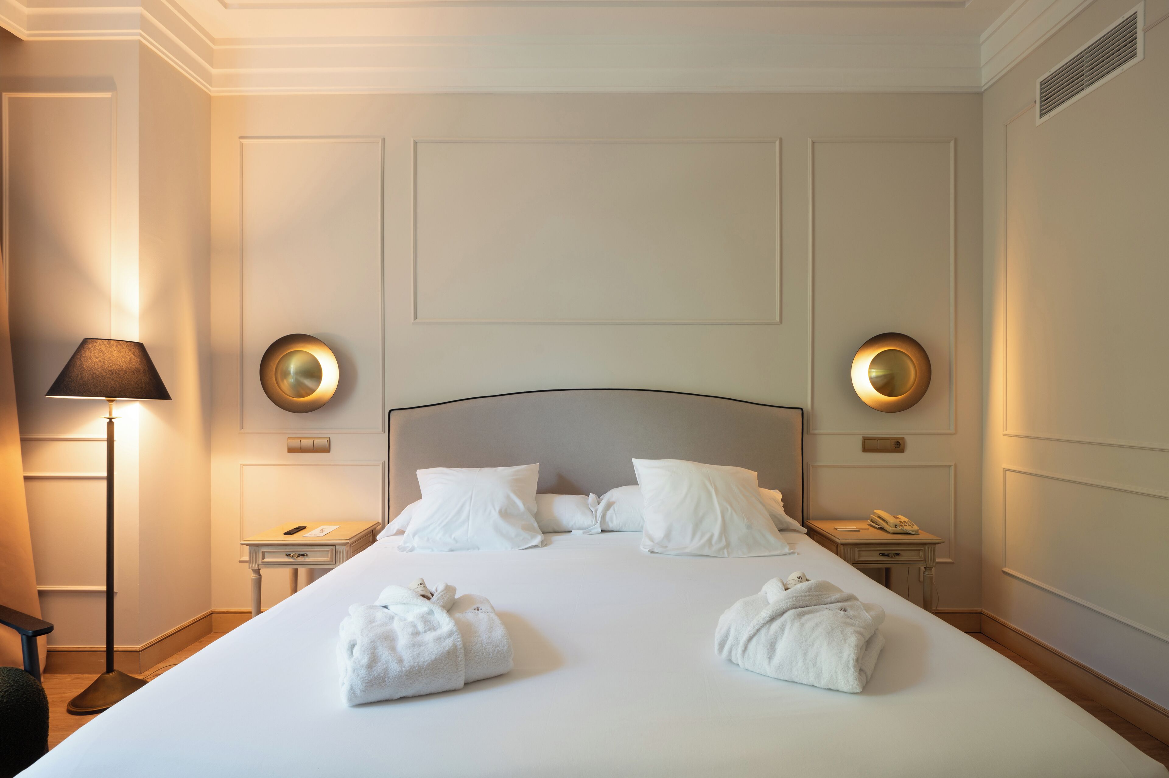 A neatly made bed with white linens and pillows, two bedside tables with lamps, and a wall-mounted light fixture.