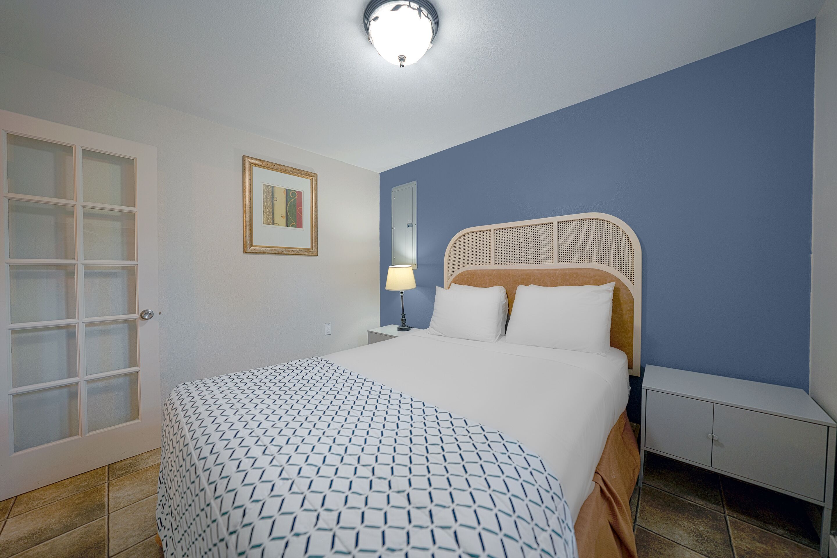 Una camera da letto con un letto, un comodino, una lampada, una porta con pannelli di vetro satinato e un quadro appeso al muro.