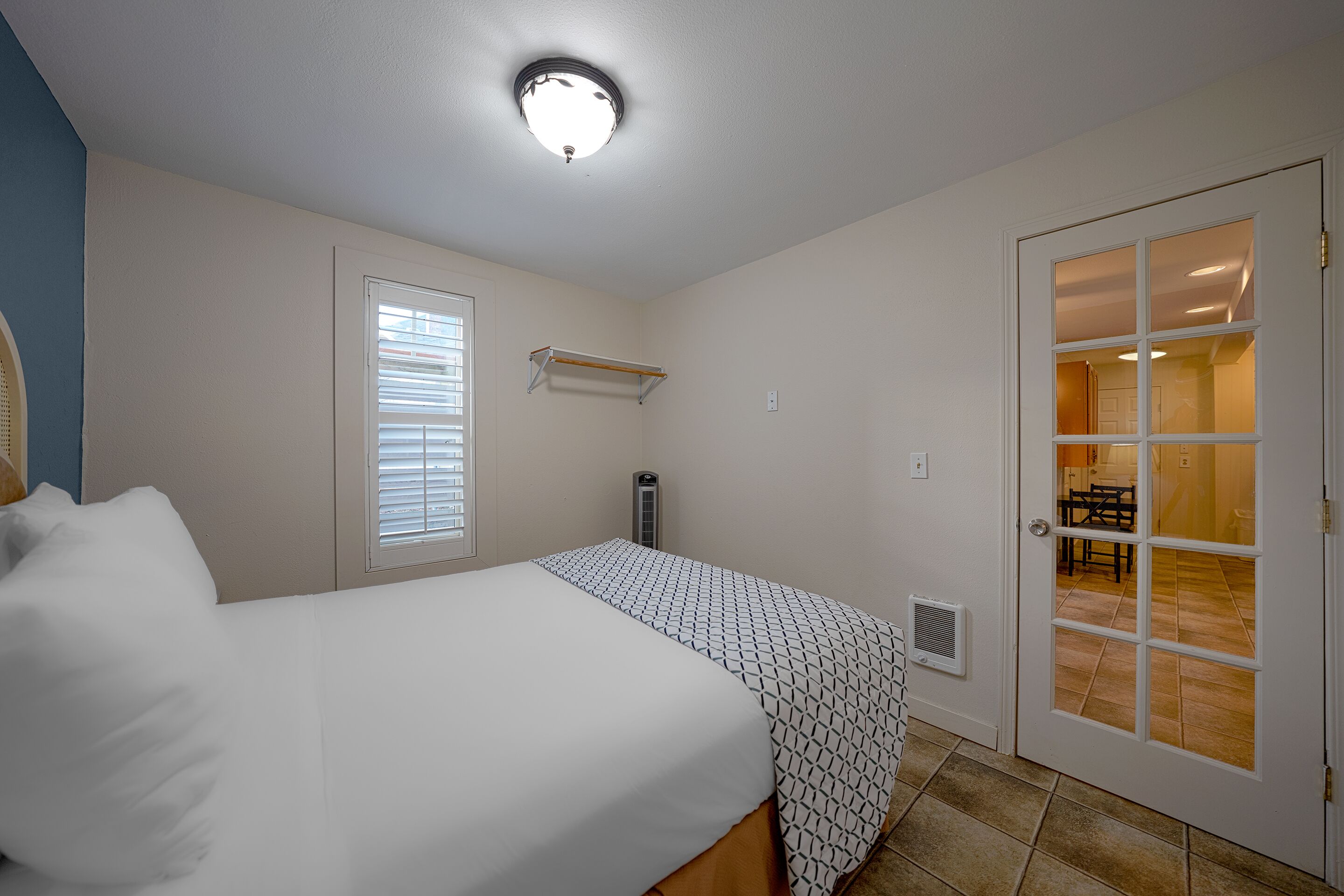 A bedroom with a bed, a window with blinds, a door leading to a dining area, and a ceiling light.