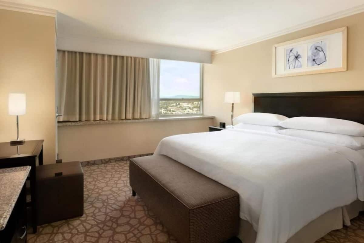 Een hotelkamer met een bed, een bankje, een bureau, een lamp, een raam met gordijnen en een ingelijst schilderij aan de muur.