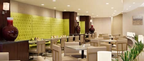 A modern hotel lobby with a green cushioned seating area, multiple tables and chairs, and decorative vases.