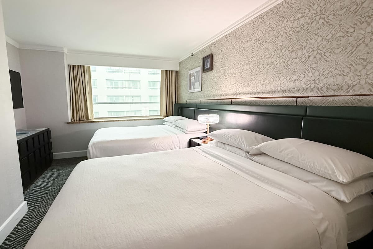Une chambre d’hôtel avec deux lits, une table de chevet avec une lampe, une télévision et une fenêtre avec des rideaux.