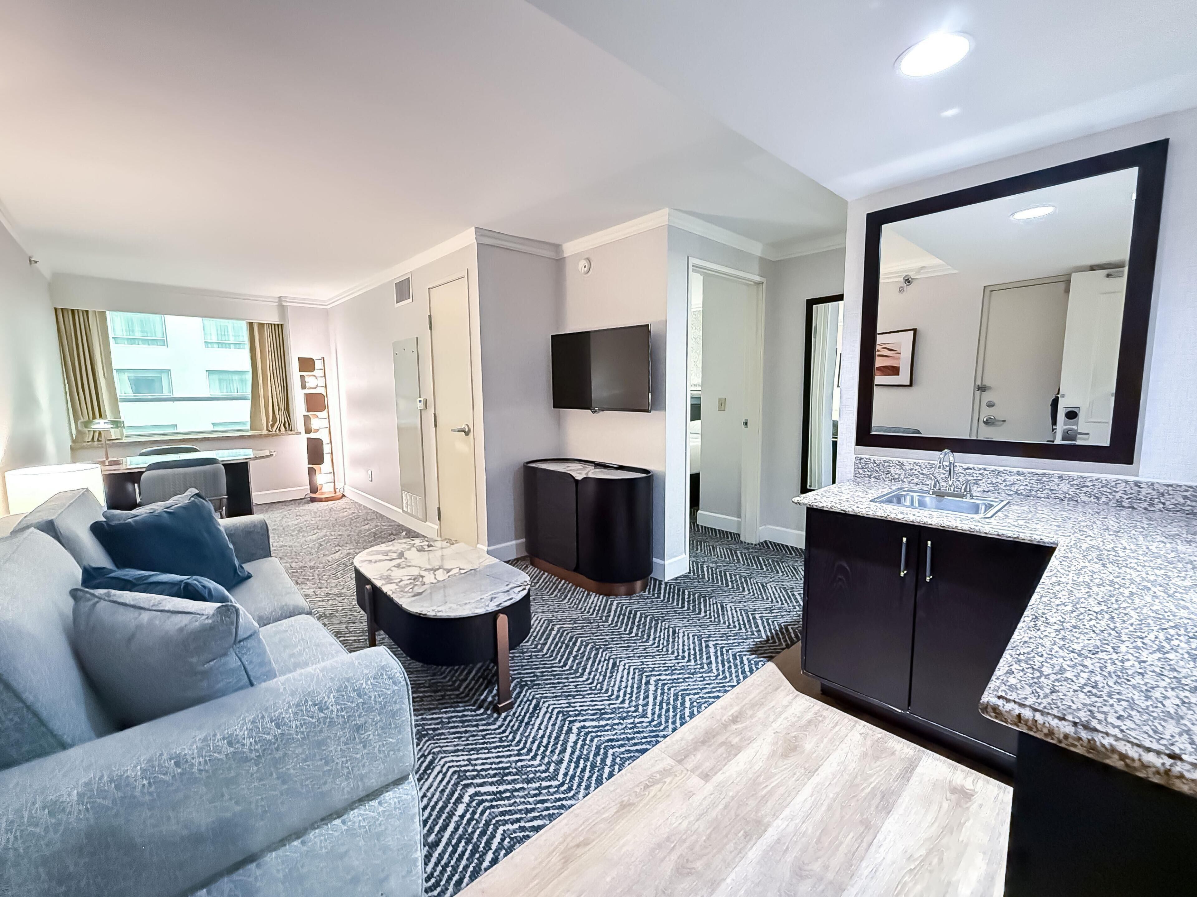 A modern hotel room with a sofa, a small round table, a TV, a mirror, and a kitchen area with a sink and countertop.