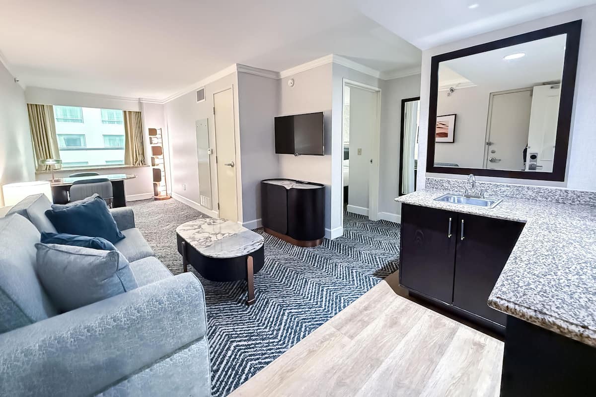 Een moderne hotelkamer met een bank, een klein rond tafeltje, een televisie, een spiegel en een kitchenette met een spoelbak en aanrecht.