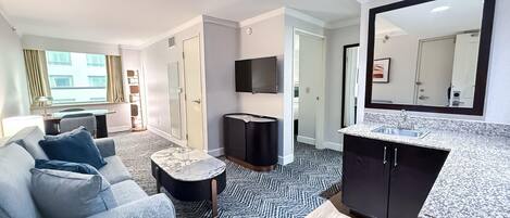 A modern hotel room with a sofa, a small round table, a TV, a mirror, and a kitchen area with a sink and countertop.