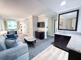 A modern hotel room with a sofa, a small round table, a TV, a mirror, and a kitchen area with a sink and countertop.