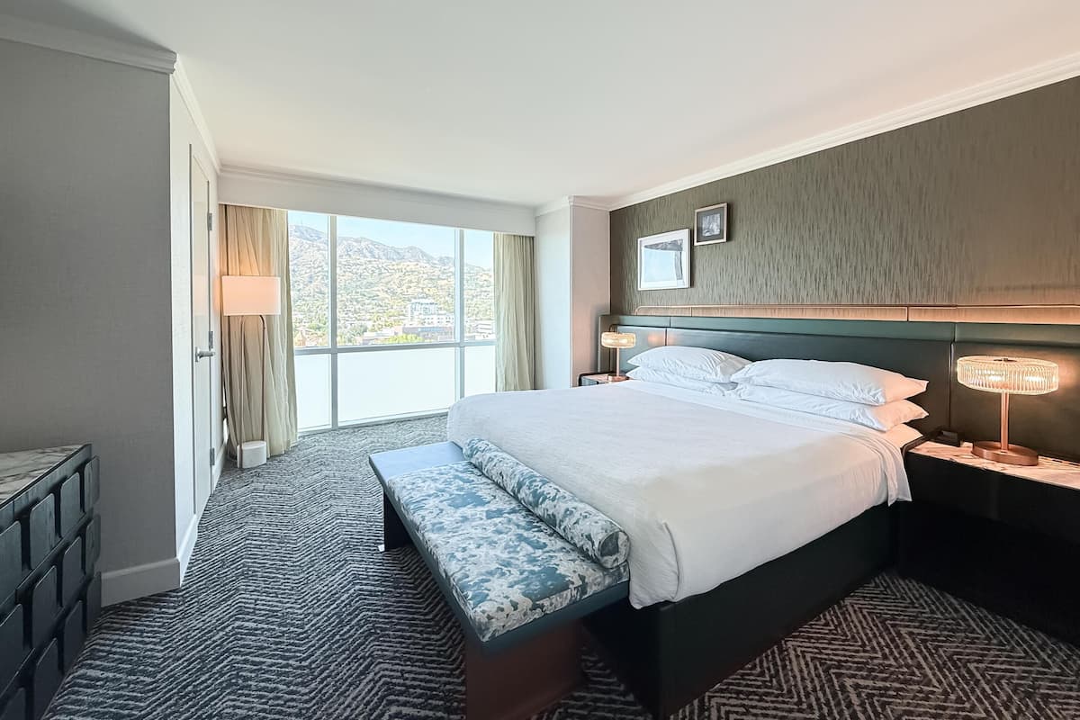 Une chambre d’hôtel avec un grand lit, une banquette, une table de chevet avec une lampe, une vue sur l’extérieur et une fenêtre avec des rideaux.