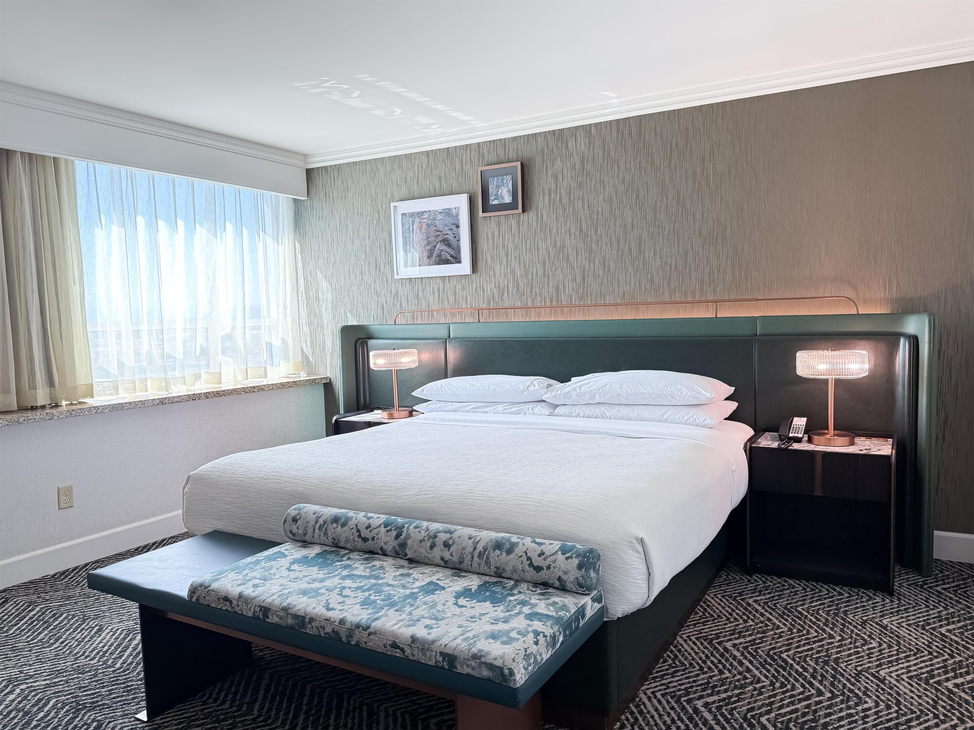 A hotel room with a bed, bedside lamps, a bench, a window with curtains, and a picture on the wall.