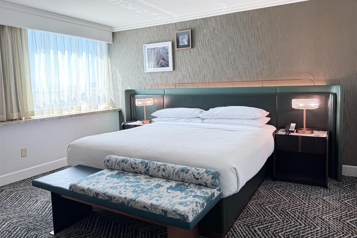 Hotelkamer met een bed, nachtlampen, een bankje, een raam met gordijnen en een foto aan de muur.